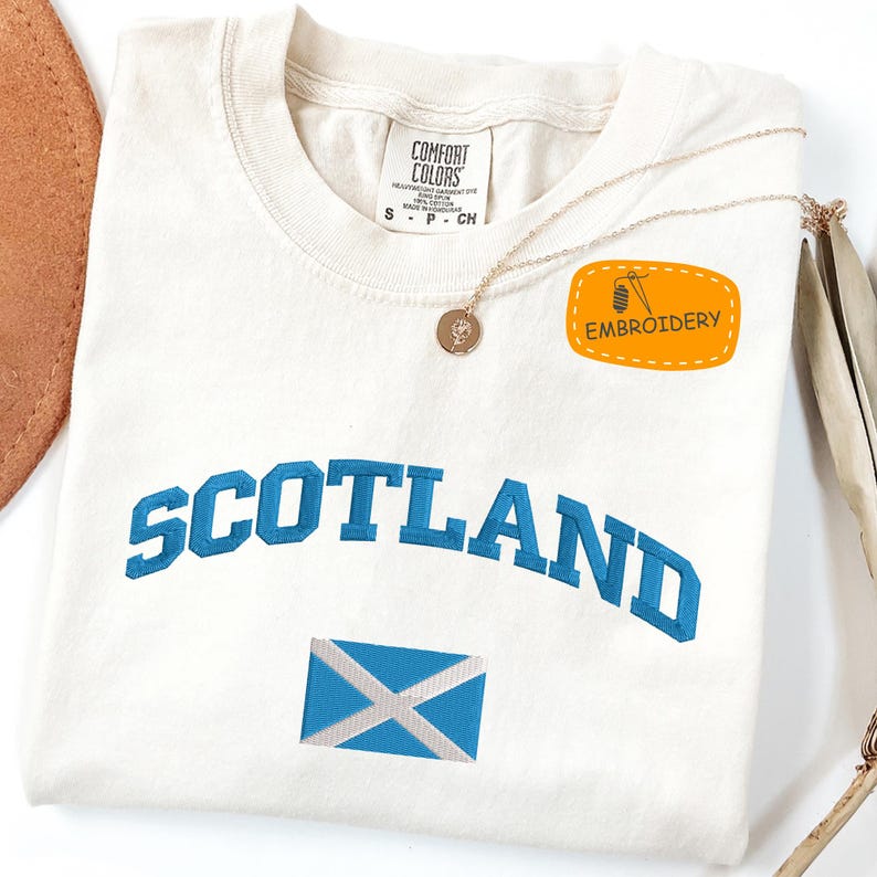 May include: Cream-colored t-shirt with "SCOTLAND" in blue lettering and a blue and white flag emblem. The shirt has a small "EMBROIDERY" patch and a gold necklace with a pendant. The shirt is made by Comfort Colors.