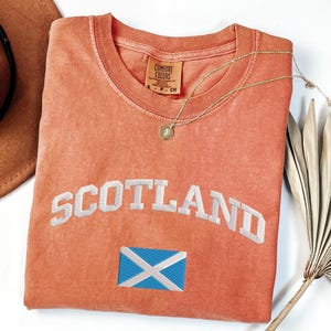 May include: A coral-colored t-shirt with the word "SCOTLAND" embroidered in white, arched across the front. Below the text is a small embroidered Scottish flag. A gold necklace with a pendant rests on the shirt. The shirt is a Comfort Colors brand.