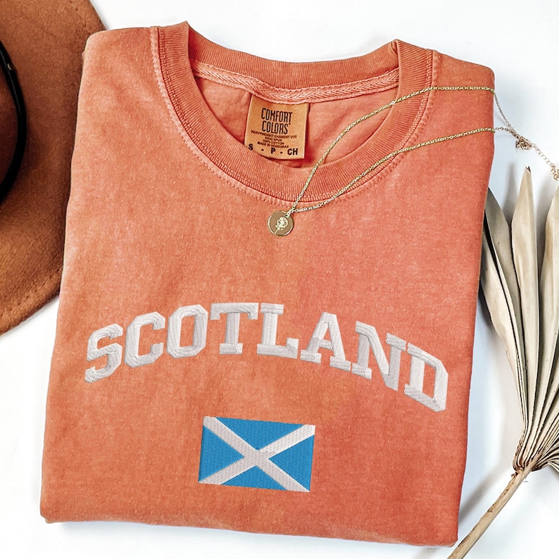 May include: A coral-colored t-shirt with the word "SCOTLAND" embroidered in white, arched across the front. Below the text is a small embroidered Scottish flag. A gold necklace with a pendant rests on the shirt. The shirt is a Comfort Colors brand.