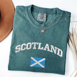 May include: Teal-colored t-shirt with the word "SCOTLAND" embroidered in white, and a blue and white flag emblem. A gold necklace with a pendant is draped on the shirt. The shirt is folded on a white surface.