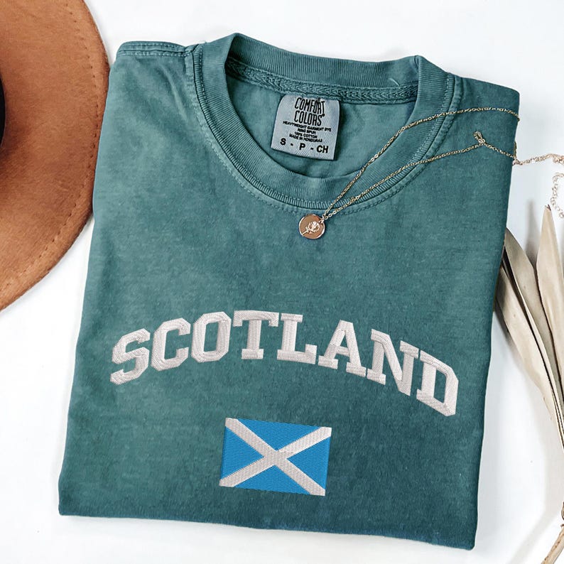 May include: Teal-colored t-shirt with the word "SCOTLAND" embroidered in white, and a blue and white flag emblem. A gold necklace with a pendant is draped on the shirt. The shirt is folded on a white surface.