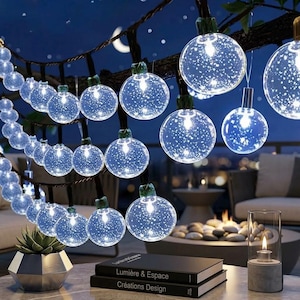 Puede incluir: Cadena de luces de globo transparente con efecto de estrellas, colgada de una rama oscura. Las luces cuelgan contra un cielo nocturno azul oscuro con una luna creciente. Una planta suculenta en una maceta geométrica y libros están en primer plano.