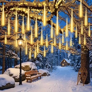 Puede incluir: Escena invernal nevada con un sendero hacia una cabaña. Luces amarillas colgantes de las ramas de los árboles simulan estrellas fugaces. Una farola negra, un banco de madera y raquetas de nieve crean un ambiente festivo y acogedor.