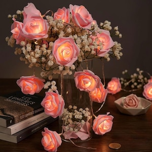 May include: Pink rose string lights with warm white illumination. The lights are arranged with a bouquet of white flowers in a clear glass vase. A small dish with a rose and books are on the table.