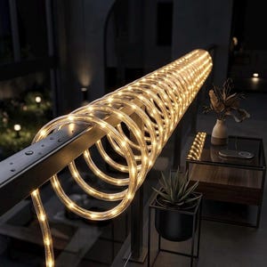 May include: A coiled string of warm white LED lights is wrapped around a black metal handrail. The lights emit a soft glow. The setting is an outdoor space at night, with a dark backdrop and a few other lights in the distance.
