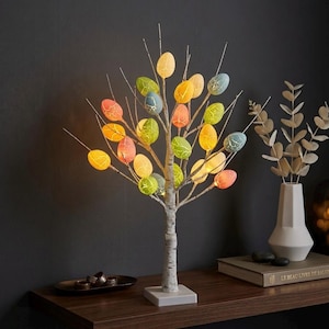 Op de afbeelding: Een decoratieve paasboom met kleurrijke, verlichte eivormige ornamenten. De boom heeft een stam en takken met berkenlook, met een vierkante witte basis. De eieren zijn in tinten geel, oranje, groen en blauw.