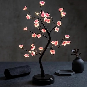 May include: A black decorative tree lamp with pink illuminated flower blossoms. The tree has a round base and a dark, textured trunk and branches. The lamp is on a dark surface with a black vase and a small black tray.