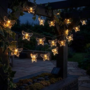 May include: A string of solar-powered butterfly lights, illuminating a garden setting. The clear butterfly lights are strung along a dark wire, with a small solar panel visible. The lights are arranged on a wooden pergola, with foliage.