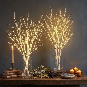 Peut inclure: Deux arrangements de branches de bouleau illuminées dans des vases en verre transparent. Les branches sont ornées de lumières LED blanc chaud, créant une douce lueur. Une bougie allumée dans un bougeoir en laiton et d'autres objets décoratifs sont sur une table en bois.