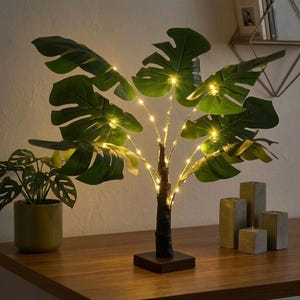 Peut inclure: Une plante artificielle de monstera avec de grandes feuilles vertes et de petites lumières LED blanc chaud. La plante a un tronc brun foncé et repose sur une base carrée en bois. D'autres objets décoratifs sont visibles en arrière-plan.