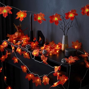Pode incluir: Uma sequência de luzes de folhas de bordo laranja com centros verdes, drapejada sobre uma superfície escura. As folhas são iluminadas, criando um brilho quente. A cena inclui abóboras decorativas, pinhas e um vaso.