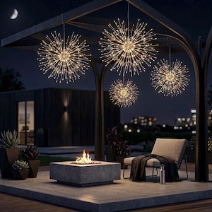 Può includere: Scena notturna all'aperto con un pergolato moderno adornato da luci a forma di stella. Un braciere quadrato con fiamme è in primo piano, con una sedia e una coperta nelle vicinanze. Sono visibili anche piante in vaso e un edificio.