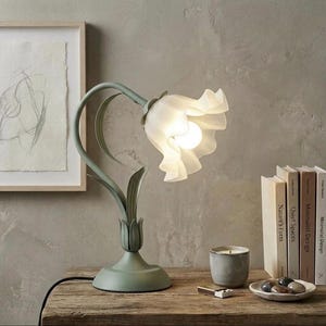 May include: A floral table lamp with a green base and stem, and a white, translucent shade. The lamp is on a wooden table, with a framed drawing, books, a candle, and a small dish. The lamp's design is reminiscent of a flower.