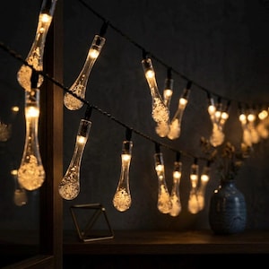 Könnte beinhalten: Lichterkette mit tropfenförmigen Leuchten, die ein warmes, gelbes Licht ausstrahlen. Jede transparente Glühbirne ist mit winzigen, funkelnden Details gefüllt. Die Lichter sind an einem schwarzen Draht befestigt und schaffen eine gemütliche Atmosphäre.