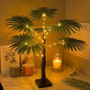 Puede incluir: Una palmera decorativa con hojas verdes y luces LED blancas cálidas. El árbol tiene un tronco marrón y se asienta sobre una base cuadrada negra. El árbol está sobre una mesa con una vela, tazas y arte enmarcado.