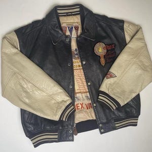 May include: Black and cream-colored leather varsity jacket with a button-up front. The jacket features cream-colored sleeves, a black body, and a collar. Patches and text are visible on the jacket, including "Avirex" and "Rex Fair Football Team".