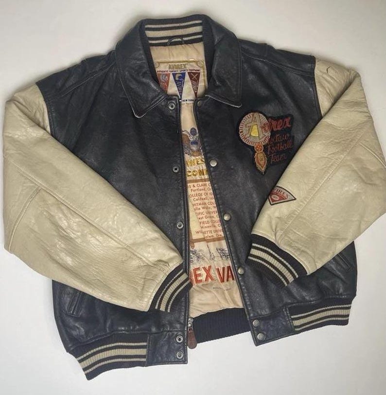 May include: Black and cream-colored leather varsity jacket with a button-up front. The jacket features cream-colored sleeves, a black body, and a collar. Patches and text are visible on the jacket, including "Avirex" and "Rex Fair Football Team".