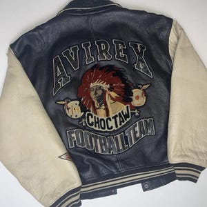 May include: A vintage black leather jacket with cream-colored sleeves. The back features an embroidered design with the word "AVIREX" above a Native American chief graphic and the words "CHOCTAW FOOTBALL TEAM".