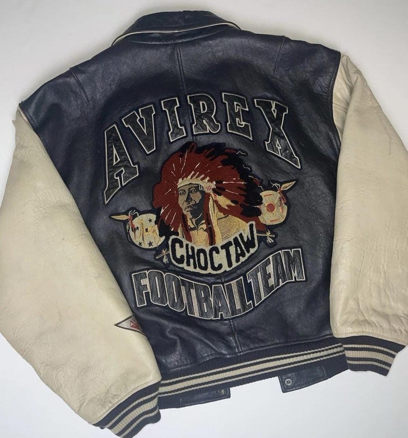 May include: A vintage black leather jacket with cream-colored sleeves. The back features an embroidered design with the word "AVIREX" above a Native American chief graphic and the words "CHOCTAW FOOTBALL TEAM".