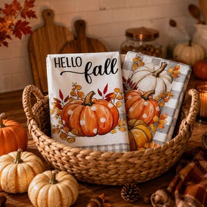 Könnte beinhalten: Zwei weiße Küchentücher, gefaltet in einem geflochtenen Korb. Ein Tuch zeigt einen großen orangefarbenen Kürbis mit weißen Punkten und den schwarzen Schriftzug "HELLO fall". Das andere Tuch hat ein graues Karomuster mit orangefarbenen und weißen Kürbissen.