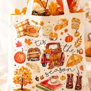 Op de afbeelding: Een crèmekleurige canvas tas met een kleurrijke herfstprint. Het ontwerp bevat een rode vrachtwagen, pompoenen, een trui, een gele paraplu, laarzen, boeken, een koffiekopje en bladeren, met de tekst 'tis the season' in zwarte schrijfletters.