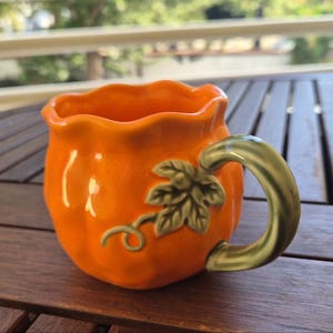 Könnte beinhalten: Ein leuchtend orangefarbener Keramikbecher in Kürbisform mit gewelltem Rand. Der Becher hat einen grünen Henkel, der wie eine Weinrebe gestaltet ist, sowie ein geformtes grünes Blatt und eine kleine, geschwungene Ranke an der Seite.