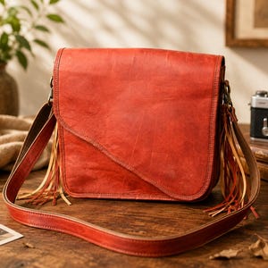 May include: A vibrant red leather crossbody bag featuring a diagonal front flap closure and long, thin leather fringe tassels hanging from both sides. The bag includes an adjustable shoulder strap and antique-style metal hardware attachments.
