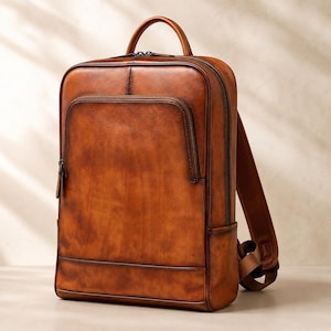 May include: A rectangular brown leather backpack with a burnished finish, featuring a top handle, a large front zippered pocket, a main zippered compartment, and adjustable shoulder straps. The design includes dark edges and visible stitching.