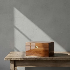 May include: A rectangular wooden box with a glossy finish and a brass keyhole. The box is a warm brown color and sits on a weathered wooden table. The background is a plain gray wall with a shadow.