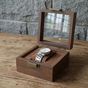 May include: A wooden watch display box with a clear glass top, open to reveal a silver wristwatch with a white face. The interior is lined with brown fabric. The box is on a wooden surface.
