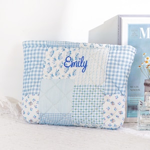 May include: A patchwork quilted pouch in shades of blue and white, with a gingham pattern, floral prints, and the name "Emily" embroidered in blue script. The rectangular pouch is likely handmade. The design incorporates various fabric patterns.