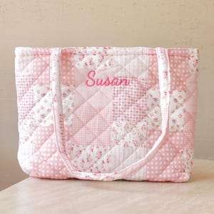 Pu&ograve; includere: Borsa tote trapuntata rosa con un design patchwork. La borsa presenta una variet&agrave; di motivi floreali e a scacchi rosa e bianchi. Il nome "Susan" &egrave; ricamato in corsivo rosa sul davanti. La borsa ha due manici.