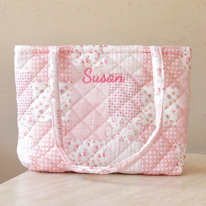 Pu&ograve; includere: Borsa tote trapuntata rosa con un design patchwork. La borsa presenta una variet&agrave; di motivi floreali e a scacchi rosa e bianchi. Il nome "Susan" &egrave; ricamato in corsivo rosa sul davanti. La borsa ha due manici.