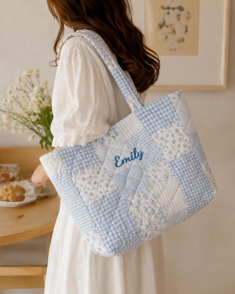 Pu&ograve; includere: Una borsa tote trapuntata con un design patchwork in blu chiaro e bianco. La borsa presenta un motivo a quadretti, stampe floreali e il nome "Emily" ricamato in blu. La borsa ha una forma rettangolare e un manico.