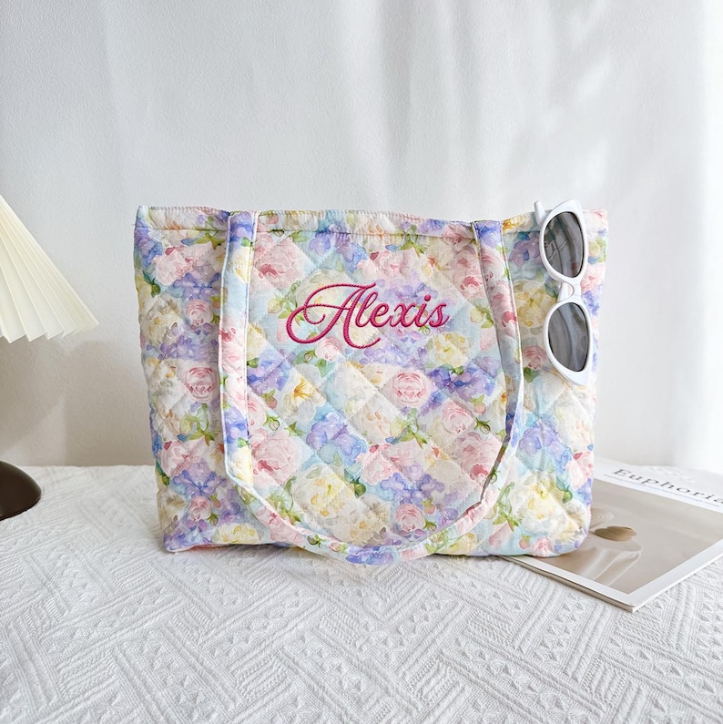 Pu&ograve; includere: Una borsa tote trapuntata con un motivo floreale in colori pastello. La borsa ha un motivo a rombi e il nome "Alexis" ricamato in rosa. Occhiali da sole bianchi sono infilati sul lato della borsa.