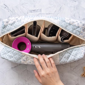 Könnte beinhalten: Eine gesteppte Stofftasche mit hellblau-weißem Karomuster, die einen dunkelgrauen Haartrockner mit einem leuchtend pinken runden Kopf sowie mehrere Haartrockneraufsätze in cremefarbenen Innenfächern aus Stoff enthält.