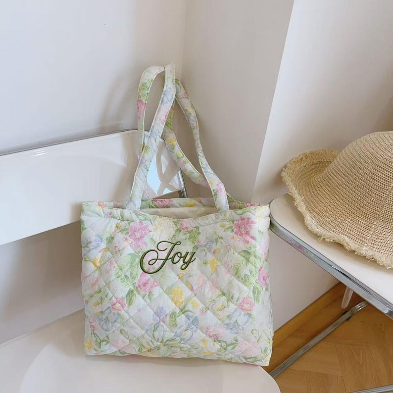 Pu&ograve; includere: Una borsa tote trapuntata con un motivo floreale in colori pastello, tra cui rosa, giallo e blu. La parola "Joy" &egrave; ricamata in verde sul davanti. La borsa ha manici coordinati e un design trapuntato a losanghe.
