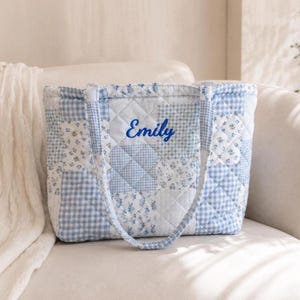 May include: A quilted tote bag with a patchwork design in shades of blue and white. The bag features a variety of patterns, including gingham and floral prints. The name "Emily" is embroidered in blue script on the front.