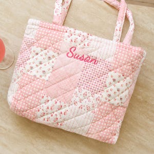 May include: A pink and white quilted tote bag with a patchwork design. The bag features floral and polka dot patterns. The name "Susan" is embroidered in pink script. The bag has matching pink handles.