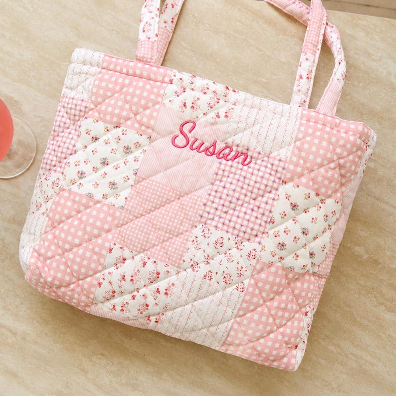 May include: A pink and white quilted tote bag with a patchwork design. The bag features floral and polka dot patterns. The name "Susan" is embroidered in pink script. The bag has matching pink handles.