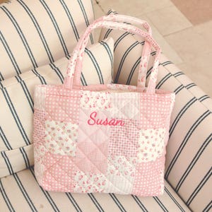 Pu&ograve; includere: Una borsa tote trapuntata rosa con un design patchwork. La borsa presenta una variet&agrave; di motivi floreali e a pois rosa. Il nome "Susan" &egrave; ricamato in corsivo rosa sul davanti. La borsa ha due manici.