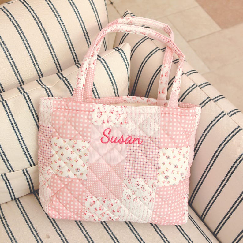 Pu&ograve; includere: Una borsa tote trapuntata rosa con un design patchwork. La borsa presenta una variet&agrave; di motivi floreali e a pois rosa. Il nome "Susan" &egrave; ricamato in corsivo rosa sul davanti. La borsa ha due manici.