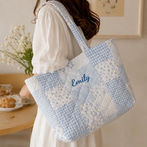 May include: A quilted tote bag with a patchwork design in shades of blue and white. The bag features a gingham pattern, floral prints, and the name "Emily" embroidered in blue. The bag has a rectangular shape and sturdy handles.