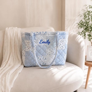 May include: A blue and white quilted tote bag with a patchwork design. The bag features floral and gingham patterns. The name "Emily" is embroidered in blue script. It has two handles and a rectangular shape. The bag is made of fabric.