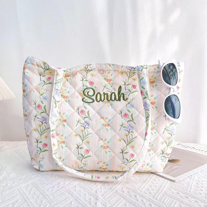 Pu&ograve; includere: Borsa tote trapuntata con motivo floreale in colori pastello. La borsa presenta un motivo a rombi e il nome "Sarah" ricamato in verde. Occhiali da sole bianchi sono agganciati al lato. La borsa &egrave; su una superficie bianca.