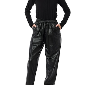 Puede incluir: Una camisa negra de manga larga con pliegues verticales y pantalones de cuero negros con cintura con cordón. Los pantalones tienen bolsillos y se combinan con zapatillas negras.