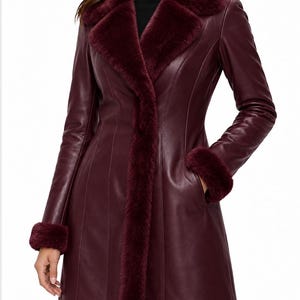 May include: A deep burgundy leather coat featuring a wide faux fur collar, matching faux fur cuffs, and vertical seam detailing. The coat has a structured fit with long sleeves and side pockets, extending to the mid-thigh area.