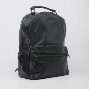 May include: Black leather backpack with a top handle and multiple zippered compartments. The backpack has a classic design with a slightly distressed finish, and features a front pocket and side pocket. The zippers are a contrasting bronze color.
