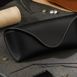 Puede incluir: Un estuche de gafas de cuero negro con costuras blancas. El estuche está rodeado de herramientas de artesanía en cuero, incluyendo hilo, agujas y un acanalador. El estuche está cerrado y tiene forma curva.