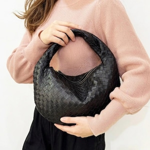 Peut inclure: Sac à main en cuir noir tressé, de forme arrondie, avec une poignée supérieure. Le sac est doté d'une fermeture éclair dorée et d'un motif tressé. Une personne portant un pull rose tient le sac.